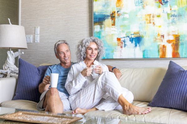 An older couple is sitting on a couch, smiling and holding mugs, with abstract artwork on the wall behind them and a lamp to their side.