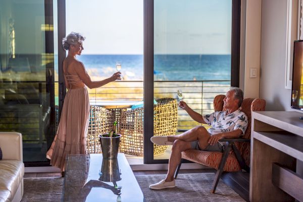 An elderly couple enjoying glasses of wine in a room with a scenic ocean view, with one seated and the other standing by the sliding door.