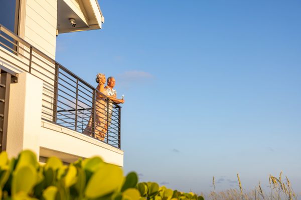 An elderly couple is standing on a balcony overlooking a serene landscape, enjoying the view together on a sunny day.