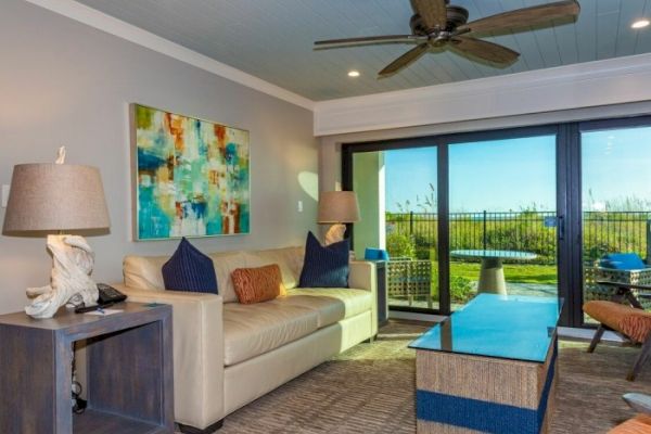 A modern living room with a beige couch, colorful artwork, a ceiling fan, and large windows showing an outdoor view and pool area ending the sentence.