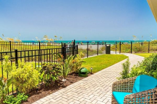 A fenced backyard with a paved pathway, plants, and outdoor seating overlooks the ocean on a clear, sunny day.