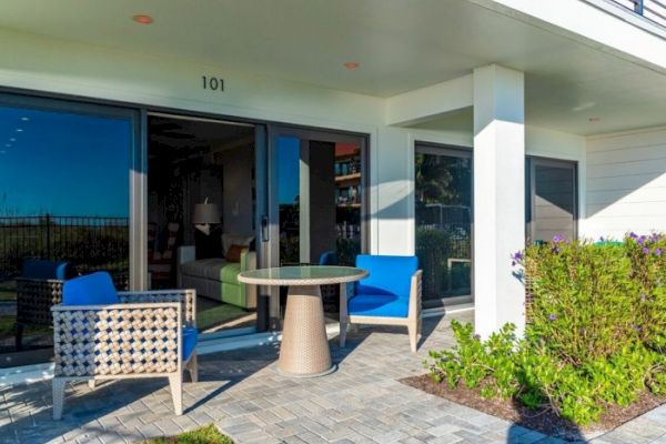 This image shows a cozy patio area with two blue chairs and a small round table outside a room labeled 101, surrounded by greenery and brick flooring.