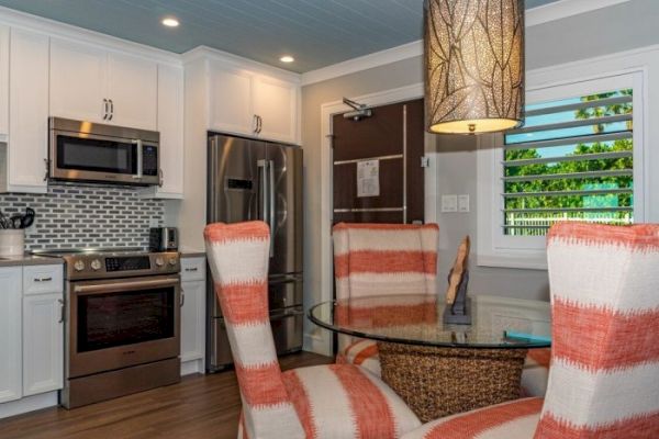 The image shows a modern kitchen and dining area with stainless steel appliances, white cabinets, a backsplash, and a table with four striped chairs.