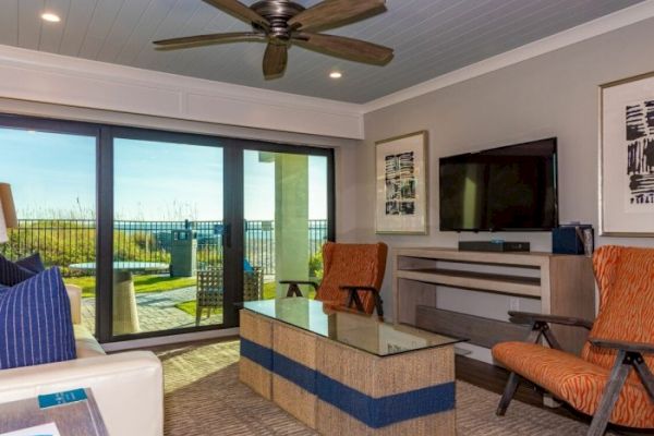 A modern living room with a large glass door, two orange chairs, a TV, and a ceiling fan, overlooking an outdoor area with a view.