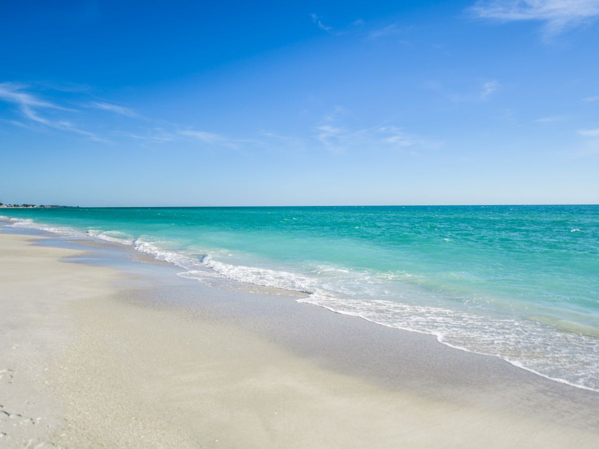 The image shows a serene beach with white sand, gentle waves, and clear turquoise water under a bright blue sky with light clouds.