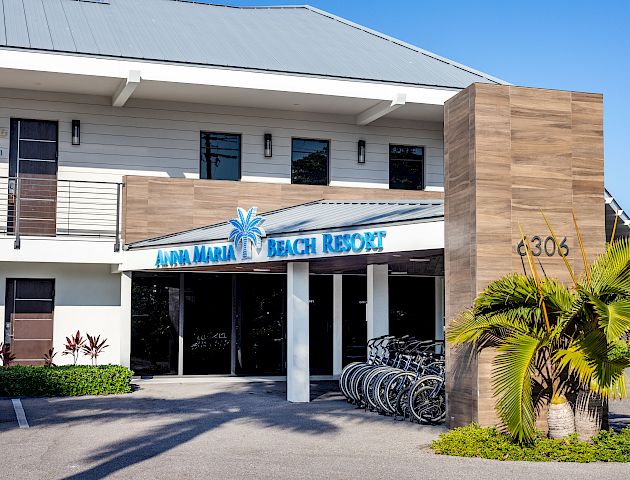 The image shows the entrance of Anna Maria Beach Resort, featuring a modern building with a sign and the address "6306," complemented by palm trees.