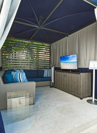 This image shows a cabana with a sofa, a flat-screen TV, a glass table, and cushioned seating, surrounded by curtains and outdoor greenery.