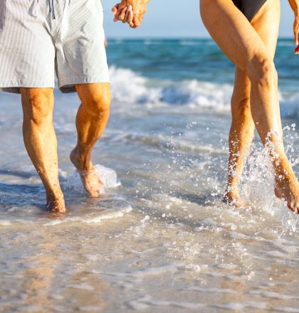 Two people are walking on the beach, their feet splashing in the water as waves crash. The ocean and blue sky are in the background.