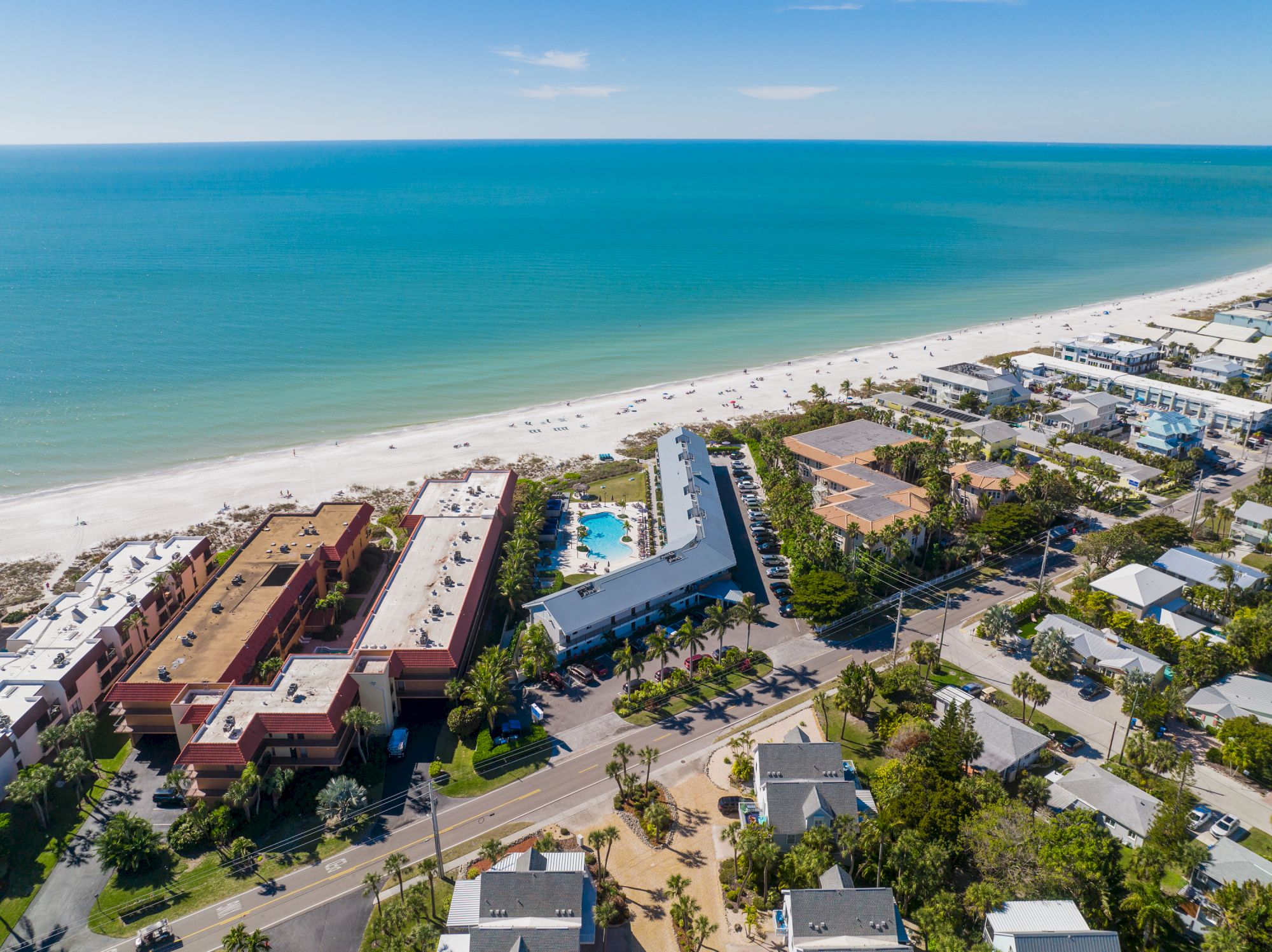 Aerial view of a coastal area featuring a beach, buildings, a pool, and surrounding residential streets ending the sentence.