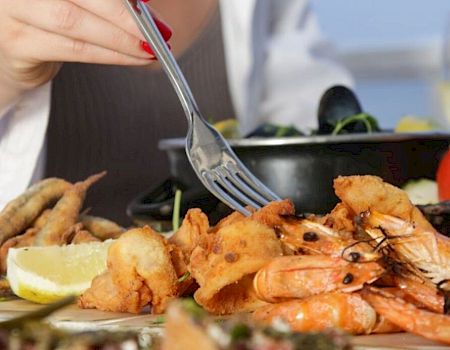 A person is dining with seafood, including shrimp, fried items, and various side dishes, on a table with a drink in the background.