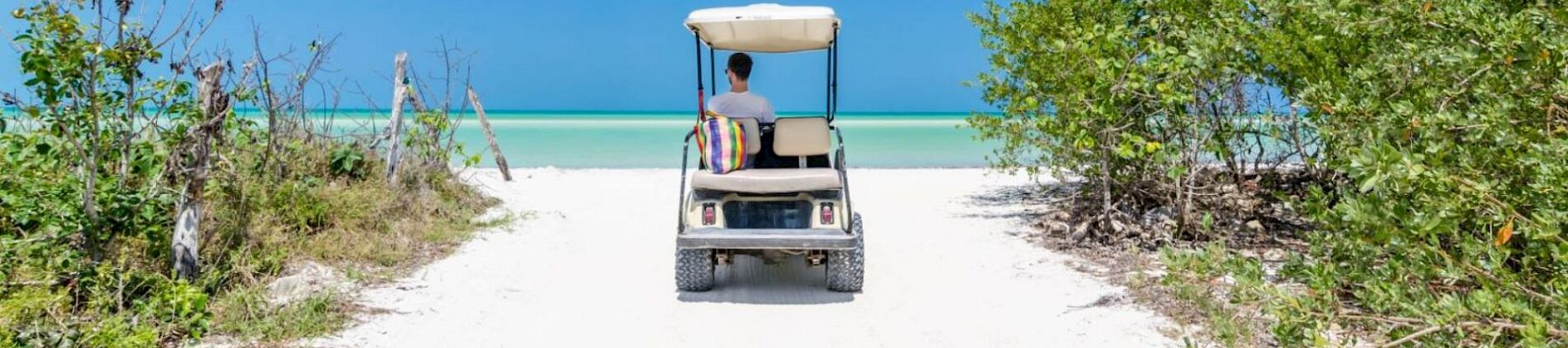 A person driving a golf cart toward a beautiful beach, surrounded by greenery, under a bright blue sky with scattered clouds.