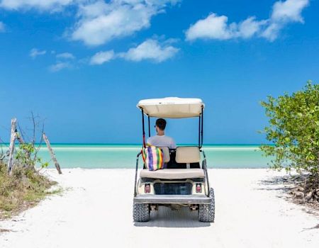 A person driving a golf cart toward a beautiful beach, surrounded by greenery, under a bright blue sky with scattered clouds.