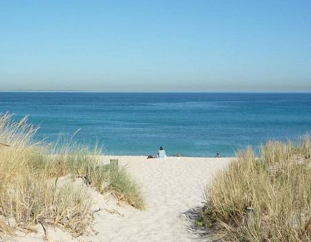 A serene beach scene with soft sand, tall grasses, and a distant view of the calm blue ocean under a clear sky.