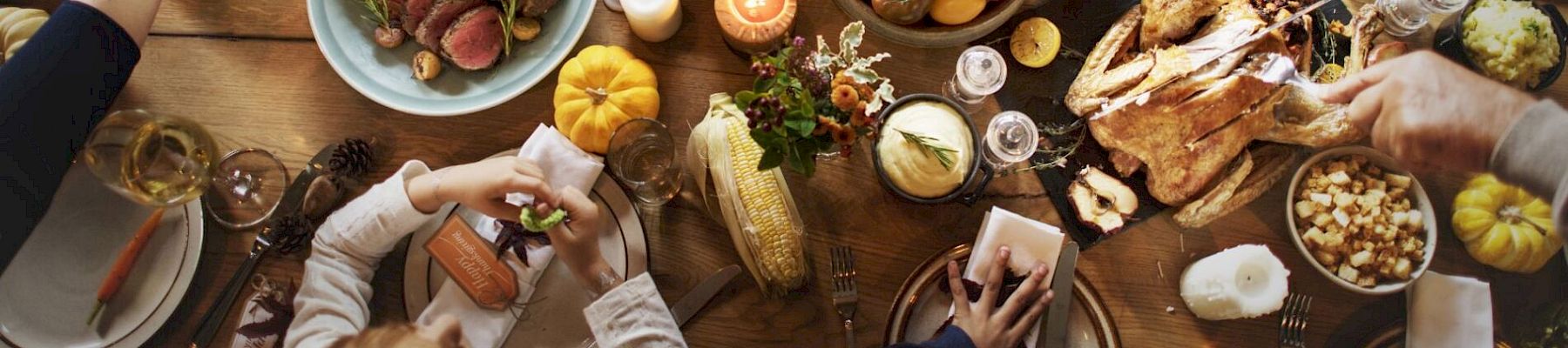 A dining table is filled with various foods: roast turkey, vegetables, fruits, and candles. People are gathered, serving, and enjoying the meal.