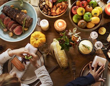 A dining table is filled with various foods: roast turkey, vegetables, fruits, and candles. People are gathered, serving, and enjoying the meal.