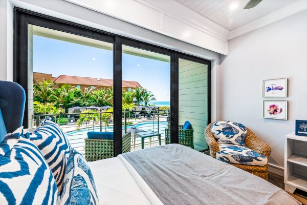 The image shows a brightly lit bedroom with a large sliding glass door leading to a balcony overlooking a pool and lush greenery.