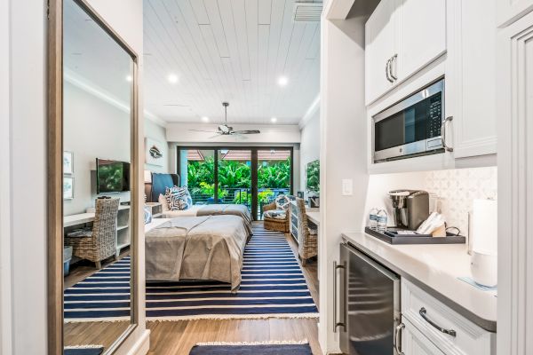 This image shows a modern, cozy bedroom with a kitchenette, featuring twin beds, a striped rug, wicker chairs, and large windows opening to lush greenery.