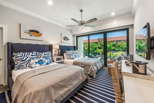 A bright, modern bedroom with two beds, tropical-themed decor, large windows with a view of lush greenery, ceiling fan, and wall-mounted TV.