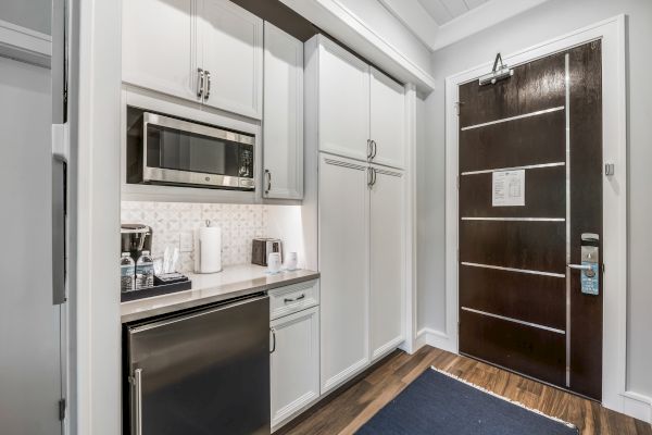 A small kitchenette with white cabinets, a microwave, a mini fridge, and a coffee maker next to a dark wooden entry door.