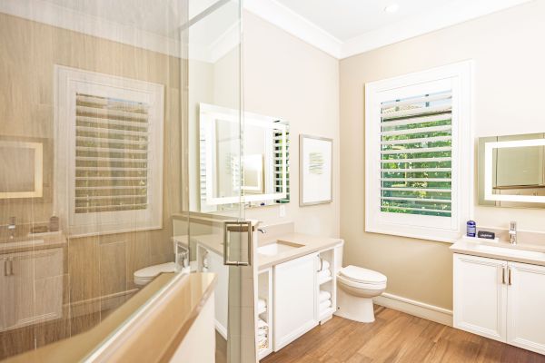 This image shows a modern bathroom with a glass shower, a toilet, a sink with a vanity, and a large mirror. The room has wooden flooring.