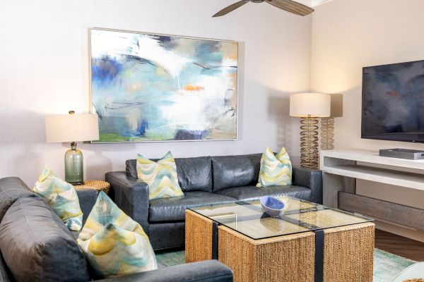 A modern living room with grey sofas, colorful throw pillows, abstract wall art, table lamps, a glass-top coffee table, and a mounted flat-screen TV.