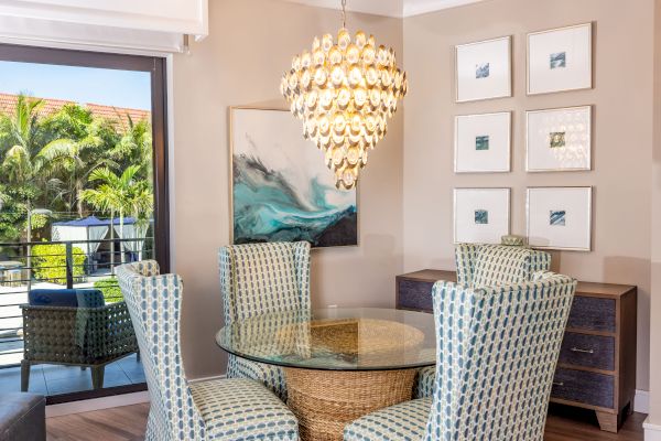 A cozy dining area features a glass table, patterned chairs, a chandelier, modern artwork, and a view of a balcony with lush greenery.