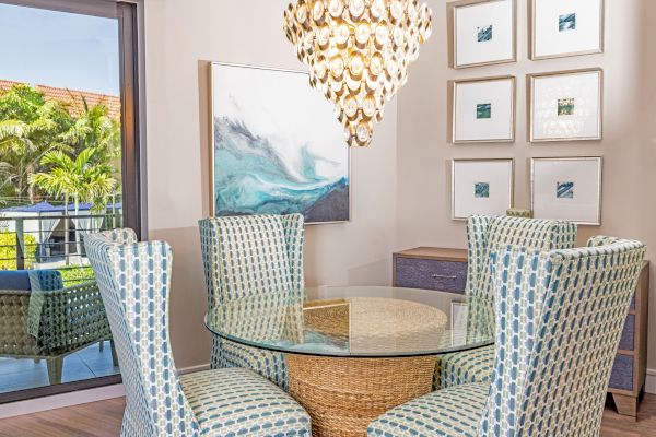 A bright dining area with a glass table, patterned chairs, a seashell chandelier, ocean artwork, and framed pictures on the wall.