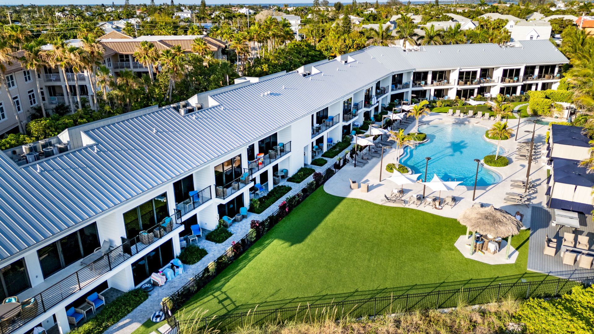 Aerial view of a resort with a large pool, sun loungers, green lawn, and surrounding buildings, amidst a lush, tropical landscape.