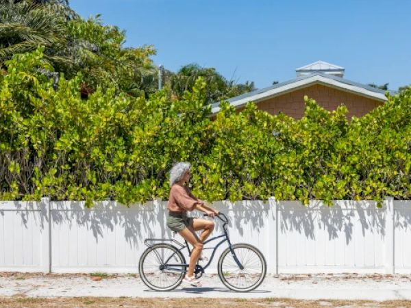 A person with grey hair is riding a bicycle along a road, passing a white fence with green shrubs and a house in the background.