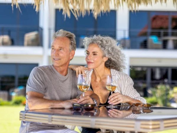 A couple is sitting at an outdoor table under a thatched roof, enjoying drinks and each other's company in a sunny setting.