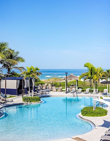 A sunny poolside at a resort with palm trees, lounge chairs, and a view of the ocean beyond, inviting and tropical.