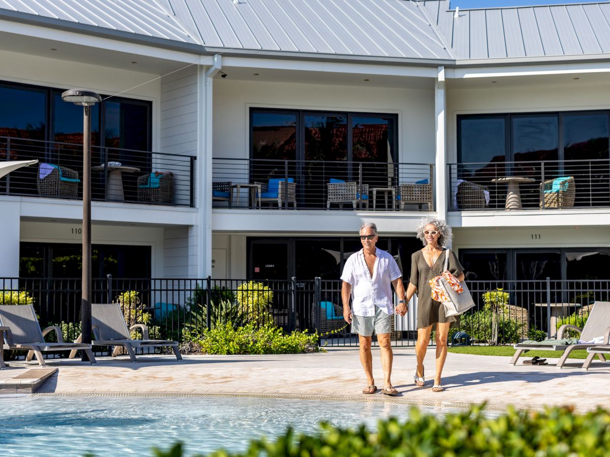 Two people stroll by a hotel pool area, with sun loungers, a modern building, and greenery in the background.