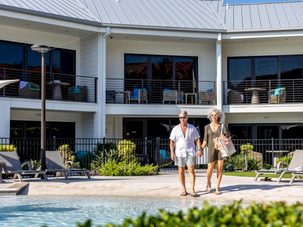Two people walk by a pool at a resort, modern white building with balconies in the background, lounge chairs, and sunny weather.